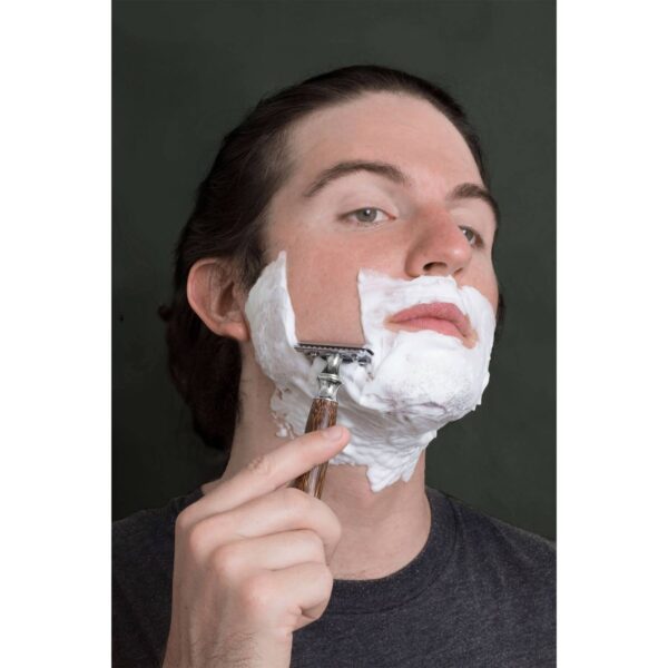 A man with shaving cream on his face looking in the mirror and shaving his face with a bamboo razor.